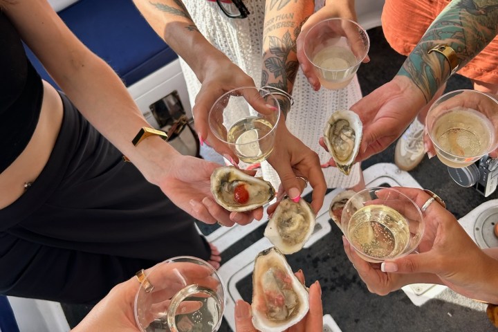 Group of people holding oysters and drinks on a boat, with visible tattoos and summer attire.