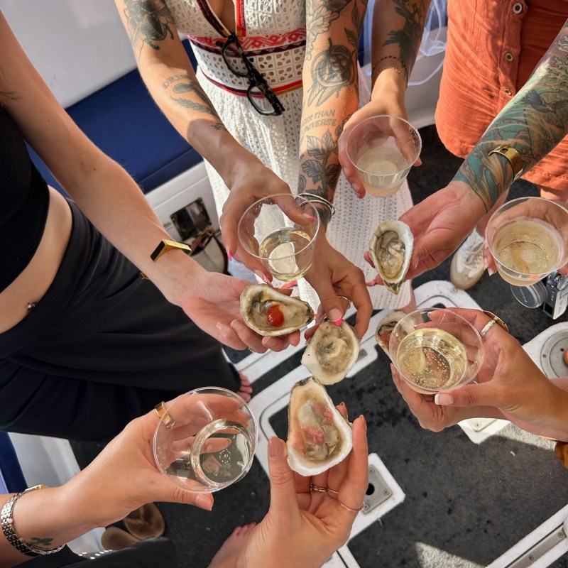 Group of people holding oysters and drinks on a boat, with visible tattoos and summer attire.