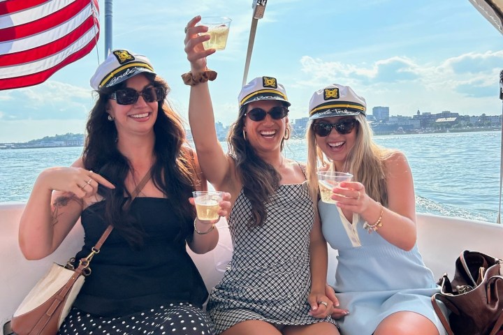 Three women in captain hats toasting on a boat with an American flag in the background.