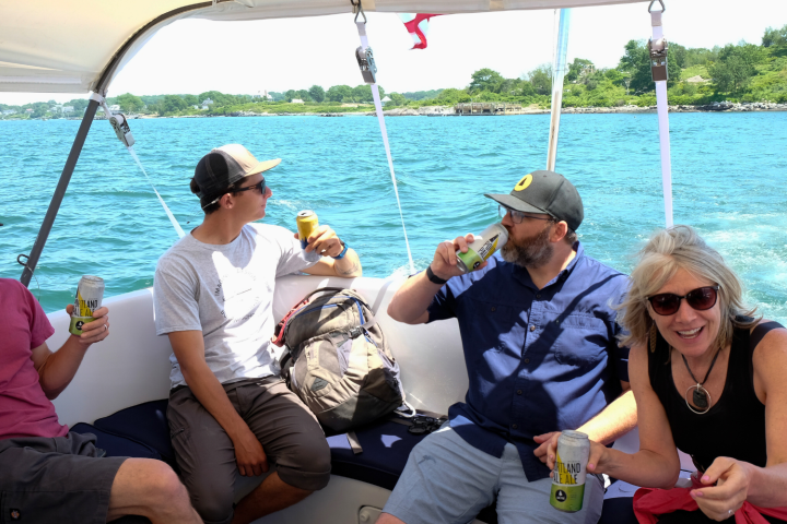 Four people on a boat enjoying drinks, set against a scenic water and shoreline view.