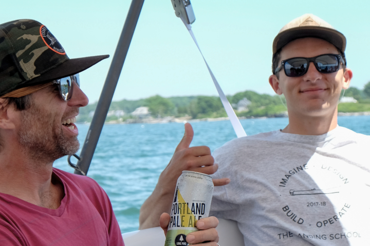 Two men in sunglasses enjoy a boat ride, one holding a drink and gesturing a thumbs-up.