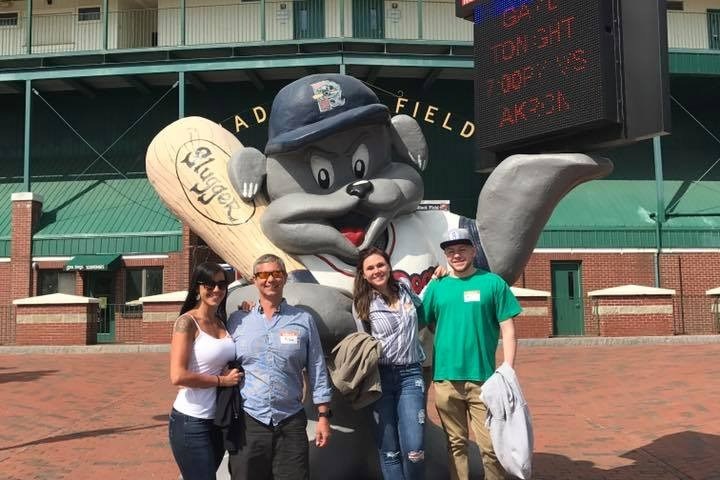 Group posing with baseball mascot statue outside stadium on a sunny day.