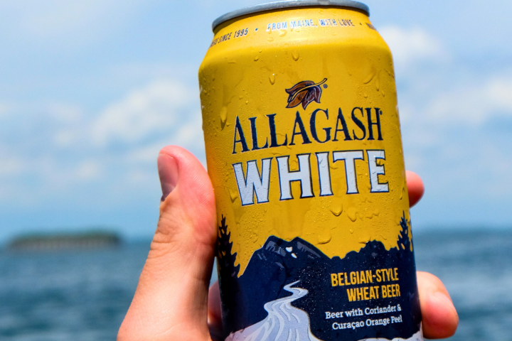 Hand holding a can of Allagash White beer with a scenic ocean background.