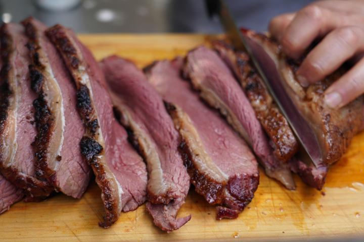 Sliced brisket on a wooden board, with a knife and hand in view.