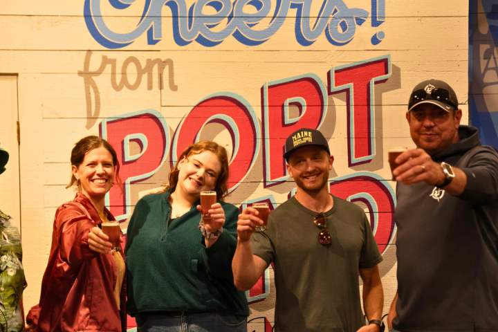 Group of five people raising glasses, smiling in front of 'Cheers from Portland' wall.