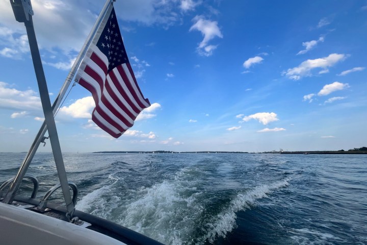 Back view from a boat with an American flag, open blue sky, and trailing wake in calm waters.
