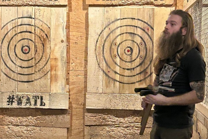 Man with long hair holding an axe in front of wooden target boards in an axe throwing venue.
