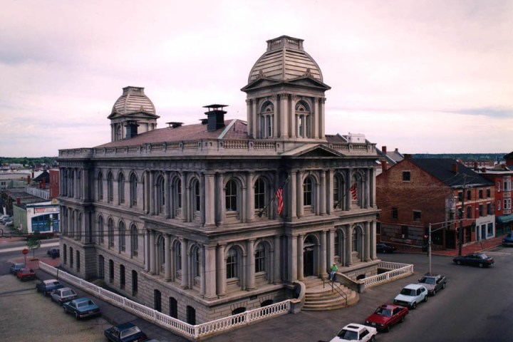 Historic three-story building with U.S. flags, dome-shaped towers, cars parked nearby, overcast sky.