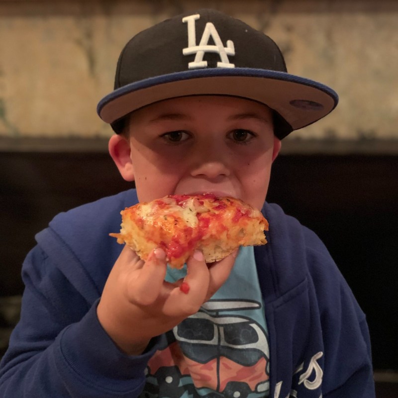 Child in a baseball cap eating a slice of pizza indoors.