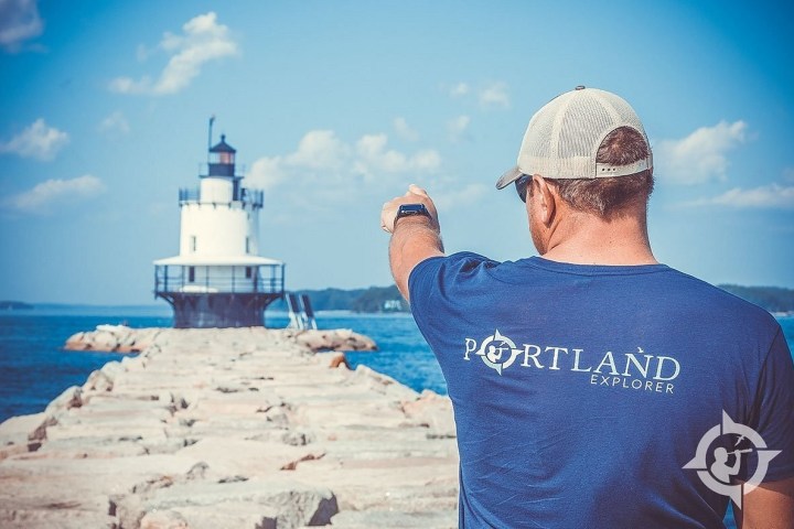 Man pointing towards a lighthouse while standing on a rocky path over water.