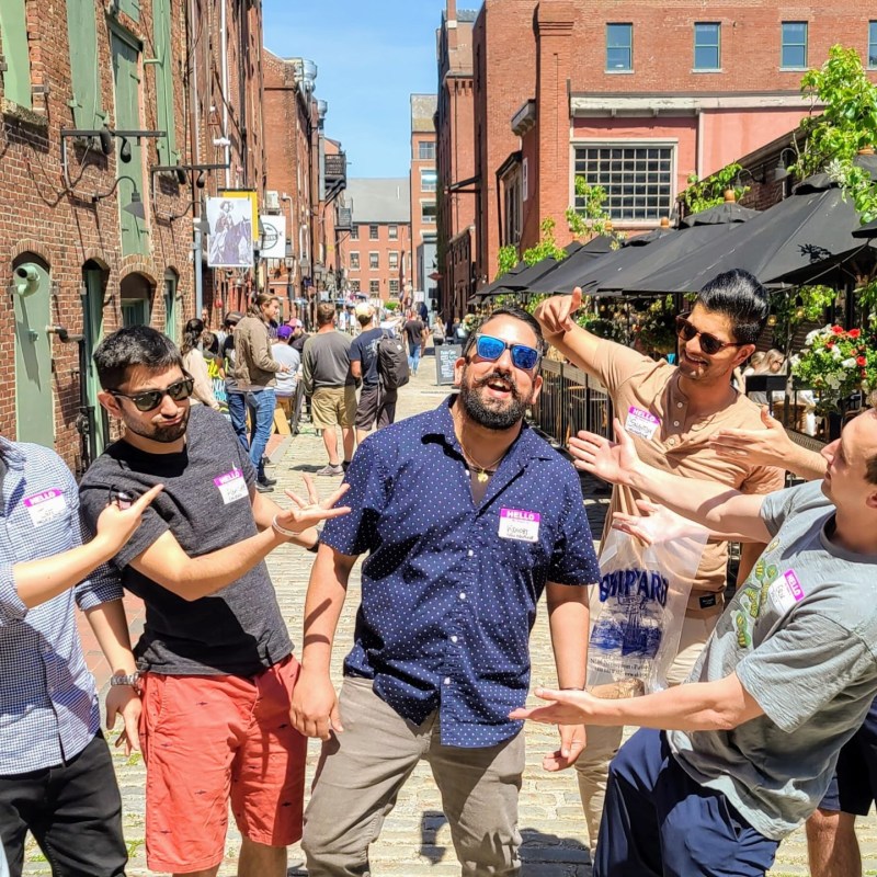 Group of men pointing at a person in sunglasses on a sunny street.