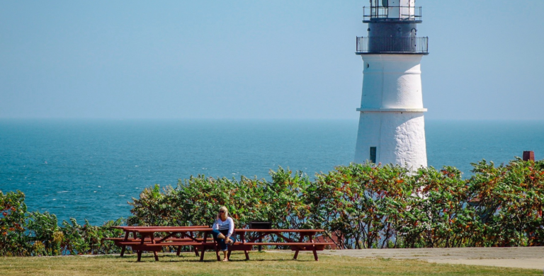 Person sitting at a picnic table near a lighthouse and ocean, surrounded by greenery.