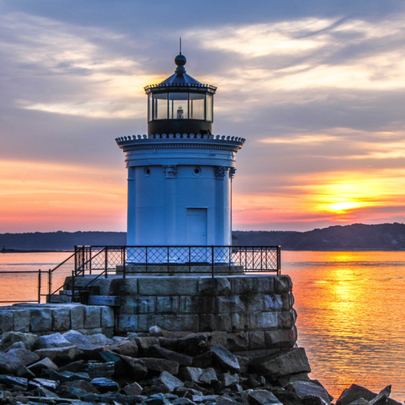 White lighthouse on rocky shore at sunset with colorful sky reflections in the water.