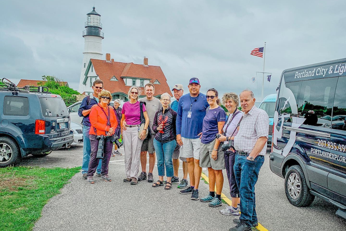 Group of people posing near a lighthouse and tour van on a cloudy day.
