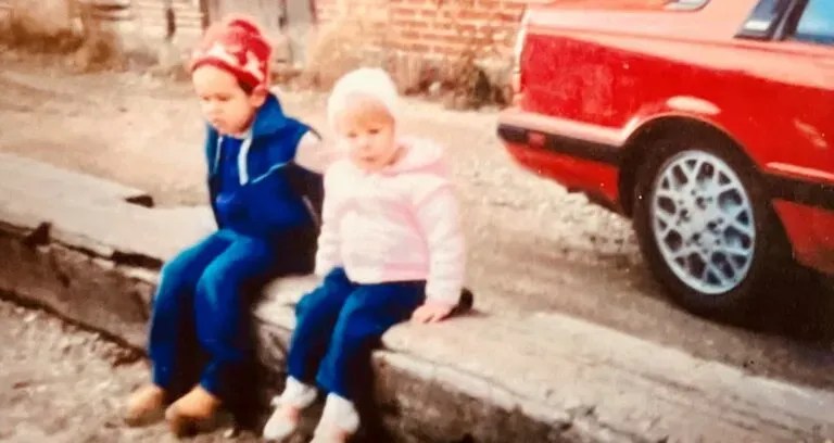 Two children in winter clothes sit on a sidewalk near a red car.