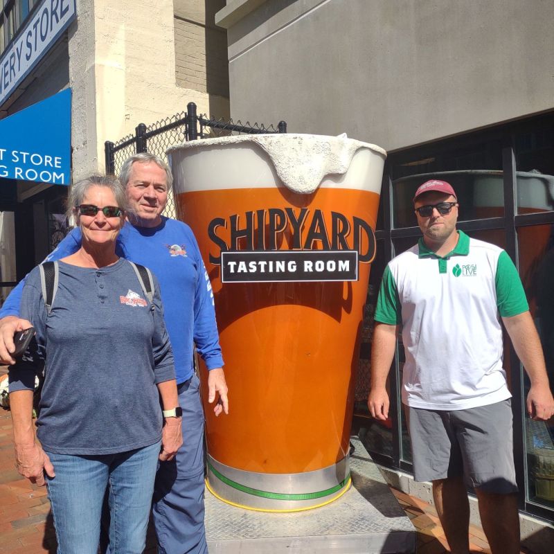 Three people standing beside a large cup with 'Shipyard Tasting Room' sign.