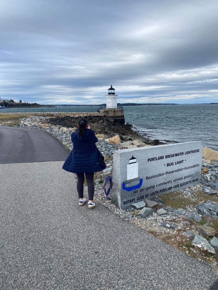 Person in coat walking toward Portland Breakwater Lighthouse by the sea on a cloudy day.