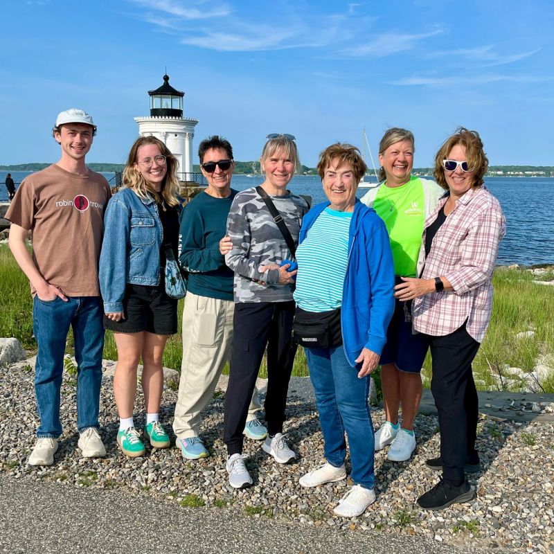 Group of seven people smiling near a lighthouse by the sea on a sunny day.