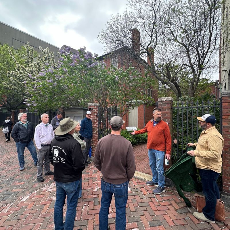 Group of people standing on a brick path near trees and a brick wall, one person gesturing.