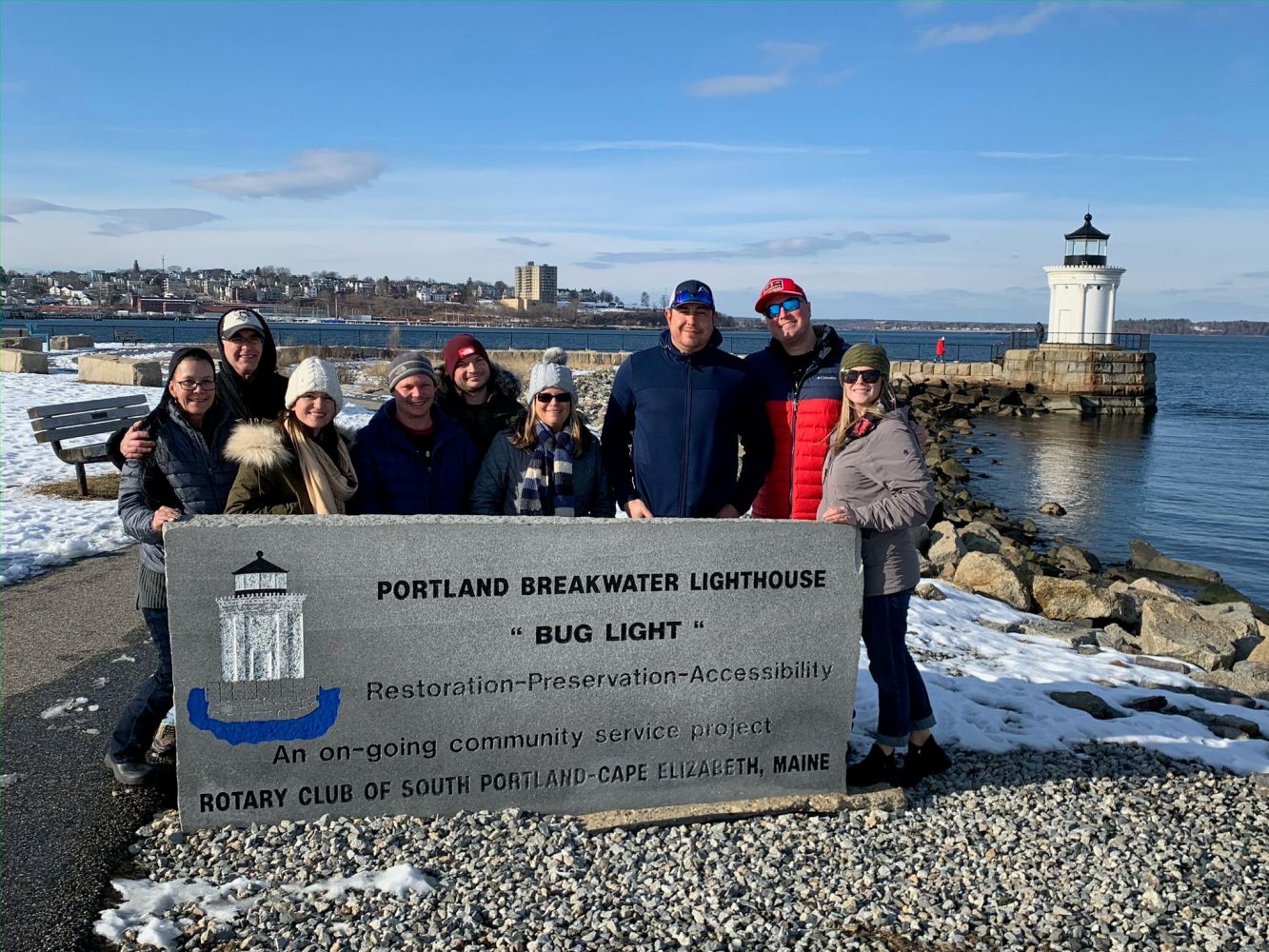Group of people posing by Portland Breakwater Lighthouse sign on a sunny winter day.