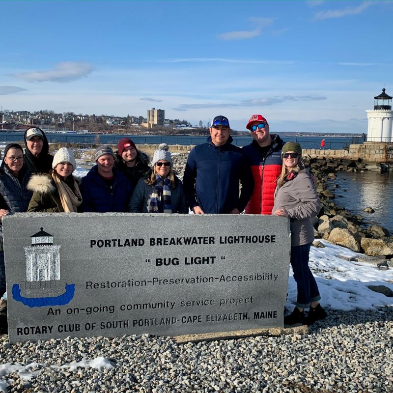 Group of people posing by Portland Breakwater Lighthouse sign on a sunny winter day.