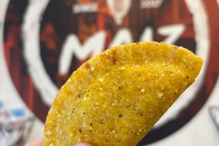 Hand holding a golden empanada with a blurred restaurant sign in the background.